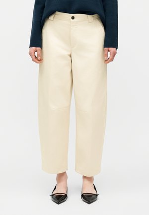 Krém színű, széles szárú, rövidített nadrágot viselő személy fekete hegyes orrú lapos cipőkkel és sötétkék hosszú ujjú felsővel, egyszínű háttér előtt állva.