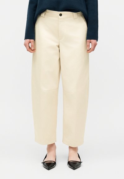 Personne portant un pantalon large crème court, des ballerines noires pointues, et un haut à manches longues bleu foncé, debout devant un fond uni.