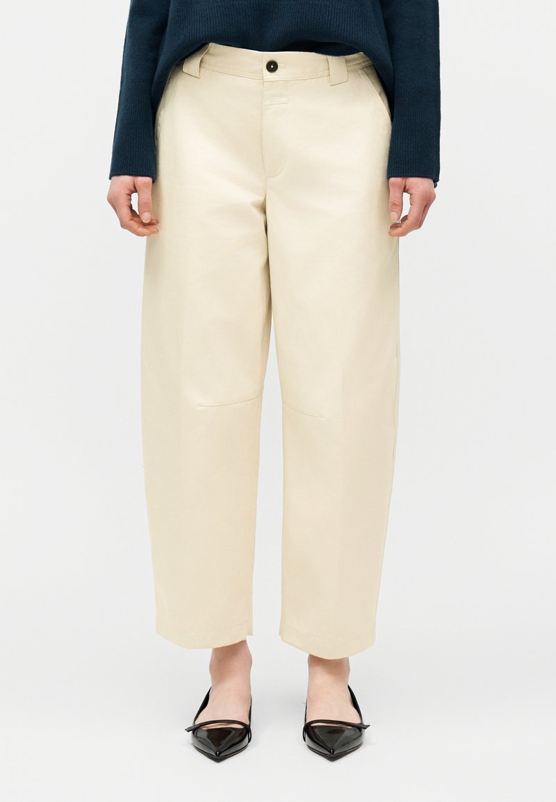 Personne portant un pantalon large crème court, des ballerines noires pointues, et un haut à manches longues bleu foncé, debout devant un fond uni.