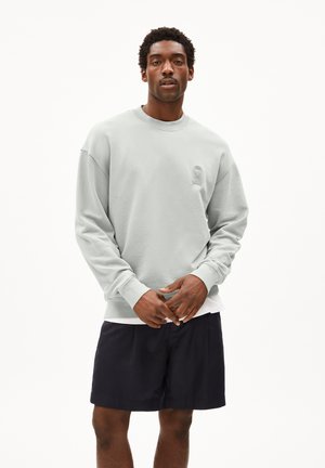 Lichtgrijze sweatshirt met ronde hals en lange mouwen, met een subtiel logo aan de voorkant. Gecombineerd met donkere shorts staat het model met de handen bijeen.