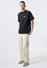 Camiseta de algodón negra con un estampado en el pecho, combinada con pantalones de algodón beige y zapatillas grises. Ajuste simple y relajado con mangas cortas.