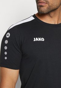 Zwarte t-shirt met korte mouwen met witte "JAKO" tekst, contrasterende witte schouderstrepen en gestippelde accenten op de mouw. Zachte stofstructuur.