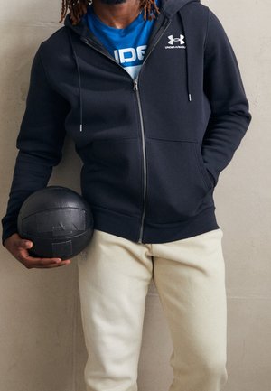 Mann trägt marineblauen Kapuzenpullover mit Reißverschluss und cremefarbene Hose, hält einen schwarzen Medizinball vor einer beigen Wand.