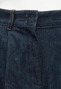 Gros plan sur la ceinture d'un jean en denim bleu foncé avec passant de ceinture visible, coutures de boutonnière et surpiqûres orange.