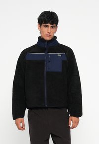 Schwarze Fleecejacke mit einem marineblauen Einsatz, Reißverschluss-Brusttasche, hohem Kragen und kontrastierendem weißen Streifen über die Brust. Lacoste-Logo auf der Tasche.
