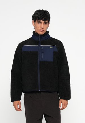 Black fleece jacket with a navy blue panel, zippered chest pocket, high collar, and contrasting white line across the chest. Lacoste logo on pocket.