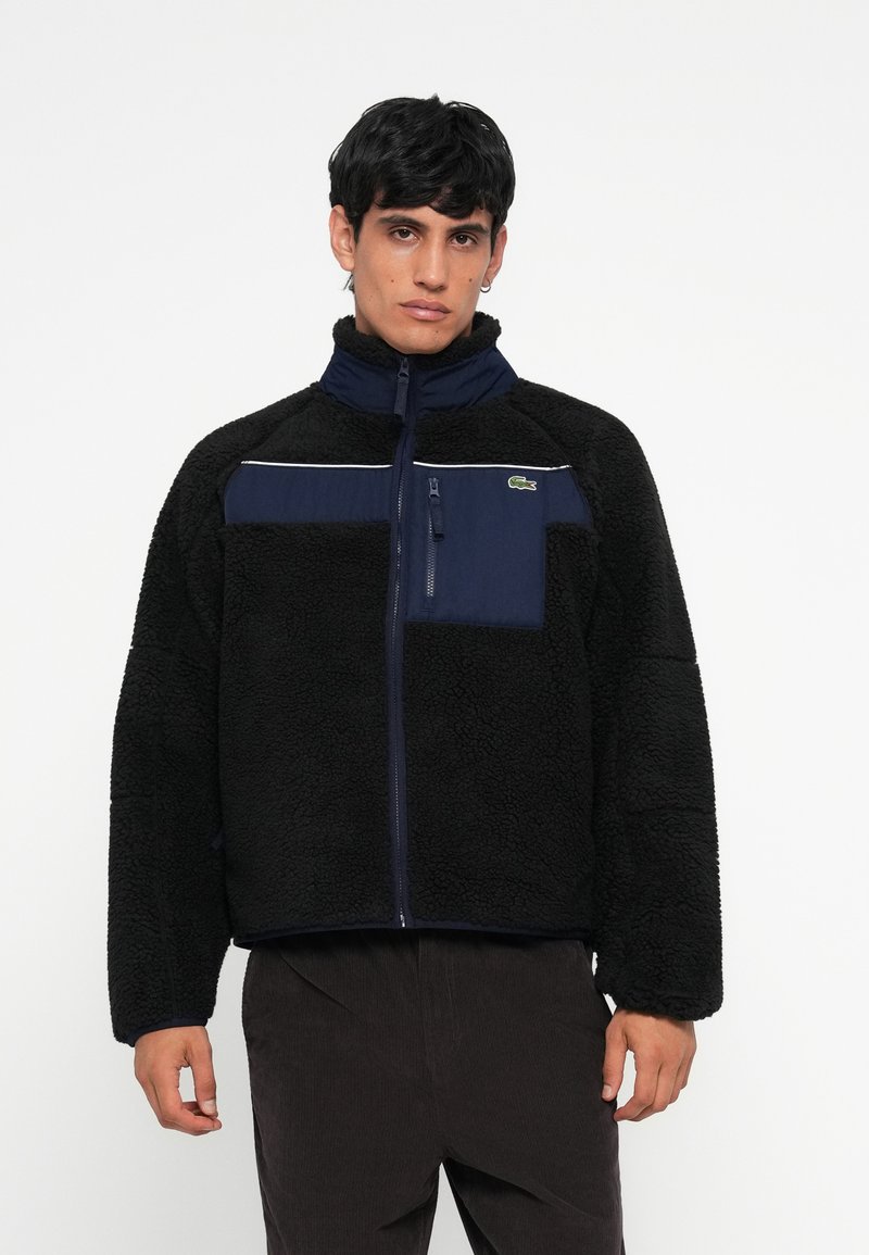 Schwarze Fleecejacke mit einem marineblauen Einsatz, Reißverschluss-Brusttasche, hohem Kragen und kontrastierendem weißen Streifen über die Brust. Lacoste-Logo auf der Tasche.