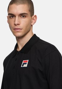 Svart polotröja med krage, prydd med en flerfärgad broderad logotyp på vänster bröst. Tyget ser slätt ut med en klassisk passform.