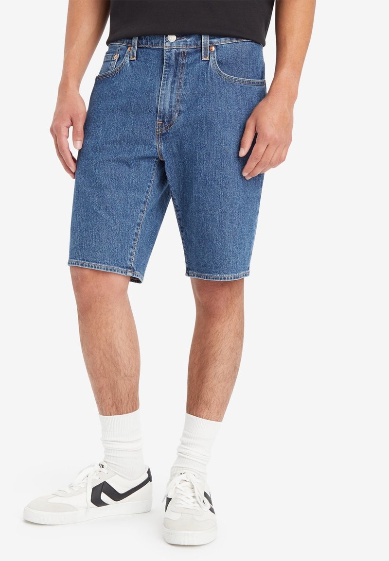 Short en denim bleu foncé, coupe droite, cinq poches, coutures apparentes ; porté avec des baskets blanches et des chaussettes.