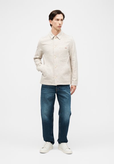 Άνδρας στέκεται με το χέρι στην τσέπη, φορώντας ένα ανοιχτό μπεζ σακάκι με κουμπιά, μπλε τζιν και λευκά αθλητικά παπούτσια, μπροστά σε απλό φόντο.