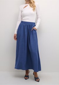 Hvid langærmet top med udskæring ved brystet, kombineret med en blå maxiskirt med elastisk talje og sidelommer.