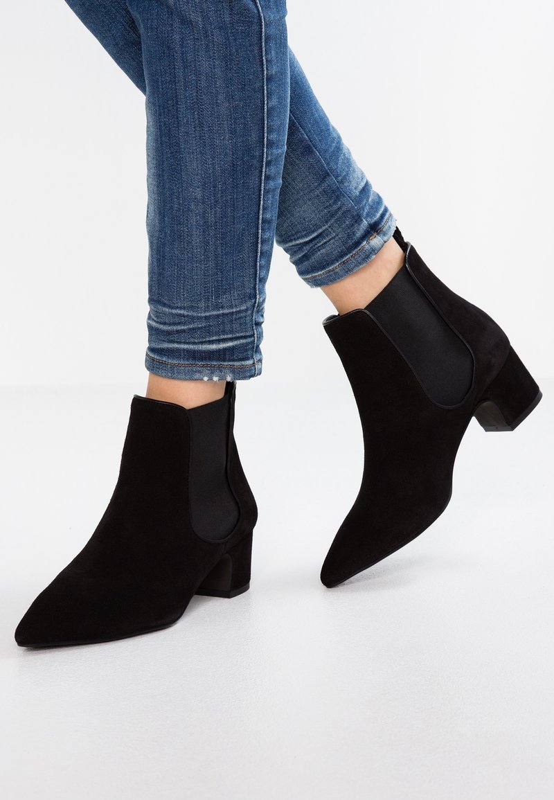 Personne portant des bottines en daim noir à talons carrés et un jean skinny bleu sur un fond blanc uni.