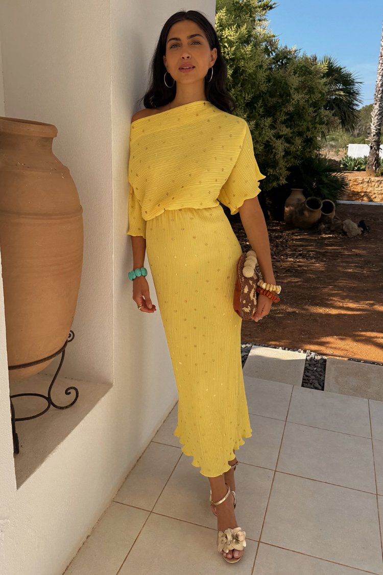 Żółta, żeberkowana sukienka midi z teksturowanym wzorem, projekt off-shoulder i zwiewnym dołem. Połączona z białymi sandałami na obcasie z kwiatowym wzorem i brązową torebką.