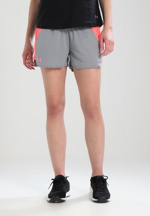 Person iført grå og koral atletikshorts, sort skjorte og sorte løbesko, der står på en ensfarvet lys baggrund.