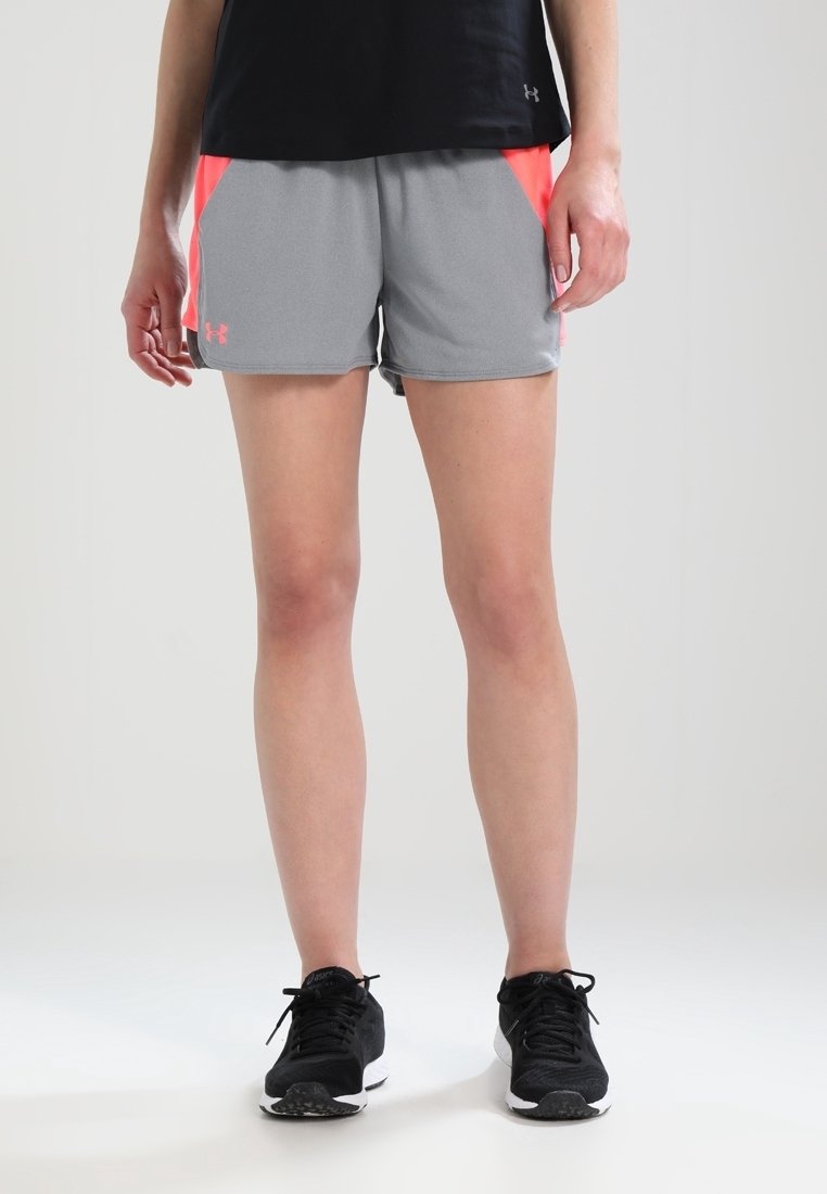 Personne portant un short de sport gris et corail, un t-shirt noir et des chaussures de course noires, debout sur un fond clair uni.