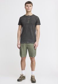 Dunkelgraues, kurzärmliges Henley-Shirt mit einer Knopfleiste aus drei Knöpfen, kombiniert mit grünen Cargo-Shorts mit Seitentaschen und braunen Wanderschuhen.