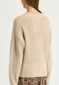 Maglione beige lavorato a maglia con trama a rete aperta, orlo a coste e spalle scese. Orecchini dorati visibili, indossati da una modella con capelli alla lunghezza delle spalle.