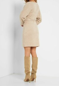 Beige klänning i stickad tröja med en figurnära midja, långa ärmar och längd till mitten av låret. Matchas med höga, slappa beige stövlar och stilettklackar.