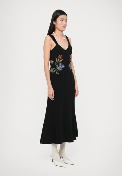 Juoda rankovių neturinti suknelė su spalvinga gėlių išsiuvinėjimu ties liemeniu, laisvo A formos silueto ir V formos iškirpte. Modelis avii baltus batus.