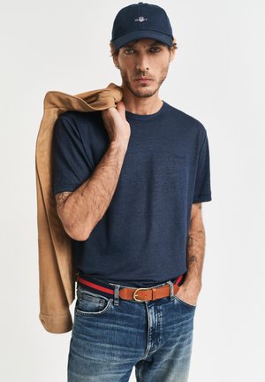 Man draagt marineblauw T-shirt en pet, houdt een beige jas over zijn schouder, met een blauwe spijkerbroek en een bruine riem, poseert tegen een effen achtergrond.