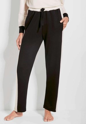 Zwarte joggingbroek met beige zijstrepen, trekkoord in de taille, voorzakken, gedragen op blote voeten door een persoon in een beige en zwarte trui.