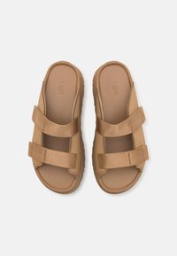 Sandales beige avec deux sangles en tissu ajustables, semelle intérieure texturée et semelle en caoutchouc épaisse. Présente le logo UGG sur la semelle intérieure.