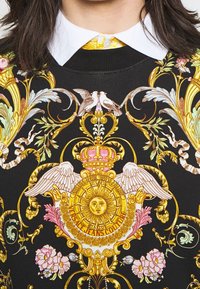 Black sweater featuring intricate gold and colorful floral patterns, with a central emblem of a sun and wings. White collared shirt underneath.