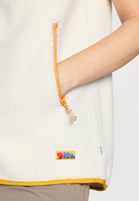 Baltas pūkuotas liemenė su oranžine užtrauktuko kišene ir geltonais akcentais. Apačioje yra išsiuvinėtas logotipas. Lygus paviršius.