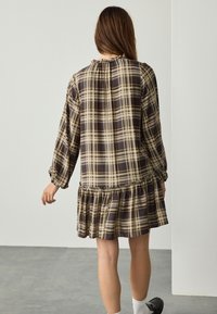 Robe à carreaux marron et beige avec manches longues, encolure froncée et ourlet évasé. Comprend un tissu texturé et des accents bleus subtils.