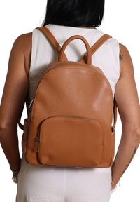 Persona che indossa uno zaino in pelle beige con tasca frontale con zip e spallacci doppi, vestita con una canotta bianca e pantaloni bianchi.
