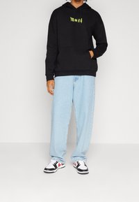 Sudadera negra con bolsillo frontal y estampado verde, combinada con vaqueros holgados de color azul claro y zapatillas en blanco y negro con acentos rojos.