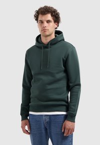 Man met krullend haar, gekleed in een donkergroene hoodie over een wit shirt en blauwe jeans, staand tegen een egale lichte achtergrond.