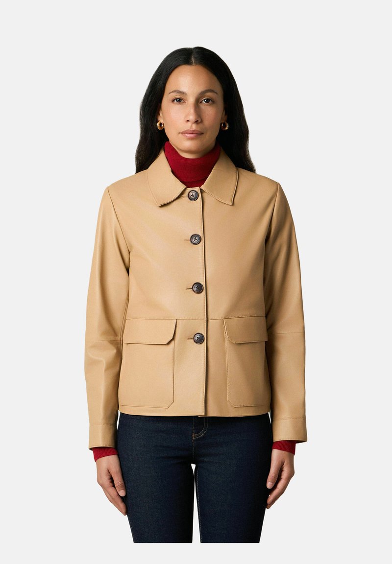 Vrouw draagt een beige leren jasje met knopen over een rode coltrui en donkerblauwe jeans, staand tegen een effen lichte achtergrond.