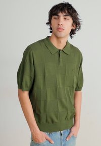 Grønn kortermet polo skjorte med et teksturert rutemønster, kragehals og ribbestrikket kant, kombinert med lyseblå denimjeans.