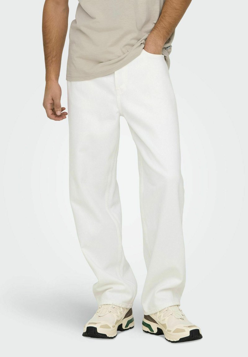 Pantalones de mezclilla blancos con un corte holgado, que presentan detalles de costura sutiles. Combinados con zapatillas beiges y verdes, complementadas por una camiseta clara.