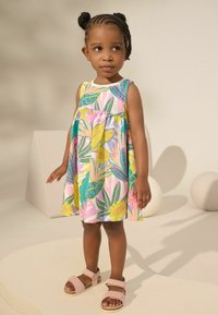 Farbiger, blumengemusterter Sommerkleid mit einem gefältelten Rock, der lebendige Grüntöne, Gelbtöne und Rosatöne zeigt. Kombiniert mit rosa Riemchensandalen.