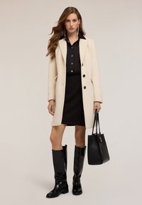Donna con capelli lunghi, che indossa un cappotto crema, un vestito nero e stivali neri fino al ginocchio, mentre tiene una borsa nera davanti a uno sfondo neutro.