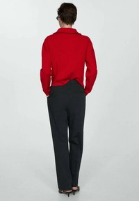 Persona con capelli corti che indossa un maglione rosso, pantaloni grigio scuro e tacchi neri, in piedi con le mani nelle tasche girata di spalle.