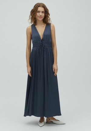 Młoda kobieta z kręconymi włosami, ubrana w granatową, bezrękawnikową suknię maxi z głębokim dekoltem w serek i białe, spiczaste baleriny.