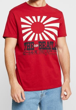 Man in een rood T-shirt met een witte opkomende zon ontwerp, de tekst "THE BEATLES", Japanse karakters en een kleine VK-vlag op de voorkant.