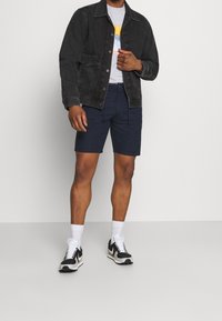 Zwarte denimjack, grijs T-shirt met opdruk, marineblauwe shorts, witte crew sokken en zwarte sport schoenen met accenten. Model staat tegen een neutrale achtergrond.