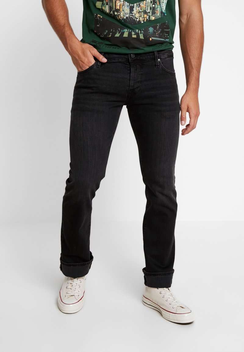 Schwarze Jeans im Slim-Fit, mit dezentem Used-Look und aufgerollten Säumen, kombiniert mit weißen Sneakern und einem dunkelgrünen T-Shirt mit Grafikdruck.