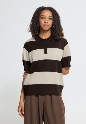 Joven mujer con cabello rizado que lleva una camiseta polo de manga corta con franjas horizontales anchas negras y beige y pantalones marrones.