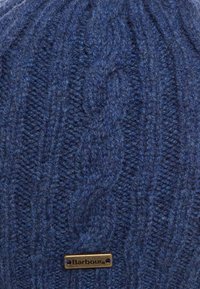 Tissu en laine tricotée bleu foncé avec motif en câble et petite étiquette métallique rectangulaire portant l'inscription "Barbour" attachée près du bas.