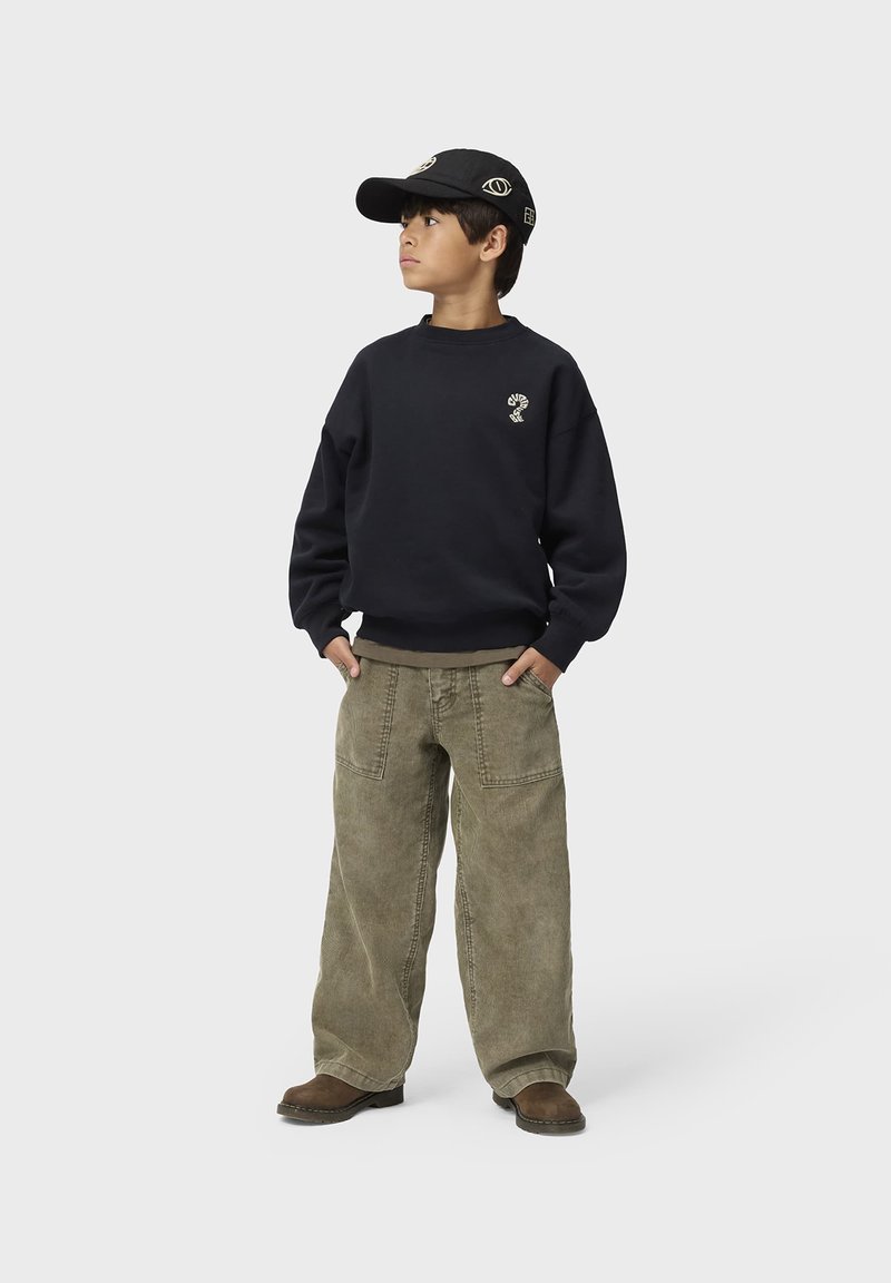 Garçon portant un sweat-shirt noir, une casquette noire avec des motifs d'yeux, un pantalon large vert olive et des chaussures marron, debout les mains dans les poches.