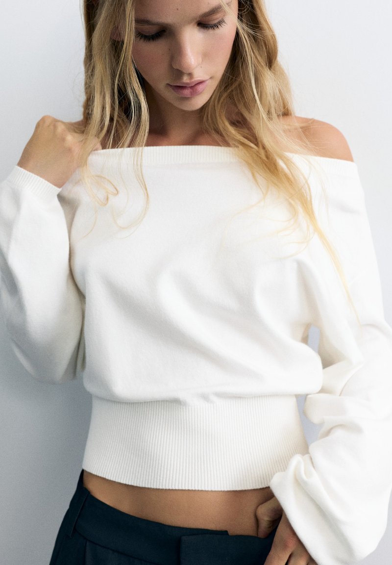 Jeune femme aux longs cheveux blonds porte un pull blanc épaules dénudées et un pantalon foncé, regardant vers le bas avec une main près du col.