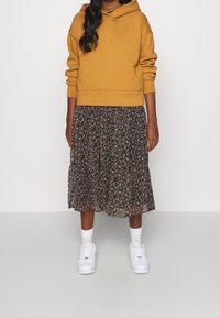 Kobieta w musztardowej bluzie z kapturem, czarnej kwiecistej spódnicy midi, białych skarpetkach i białych sneakerach, stoi na tle jasnej jednolitej ściany.