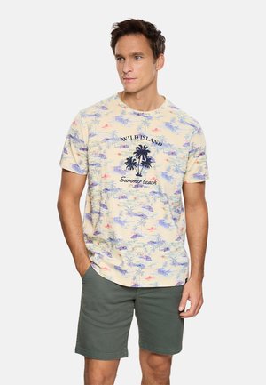 Hombre con camiseta beige de estampado tropical con texto "Wild Island Summer Beach" y pantalones cortos verde oscuro, con las manos en los bolsillos, mirando hacia la derecha.