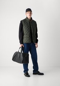 Gilet imbottito verde scuro con bottoni a pressione dorati, felpa nera, jeans blu e borsa a tracolla in pelle nera. Scarpe nere completano il look.