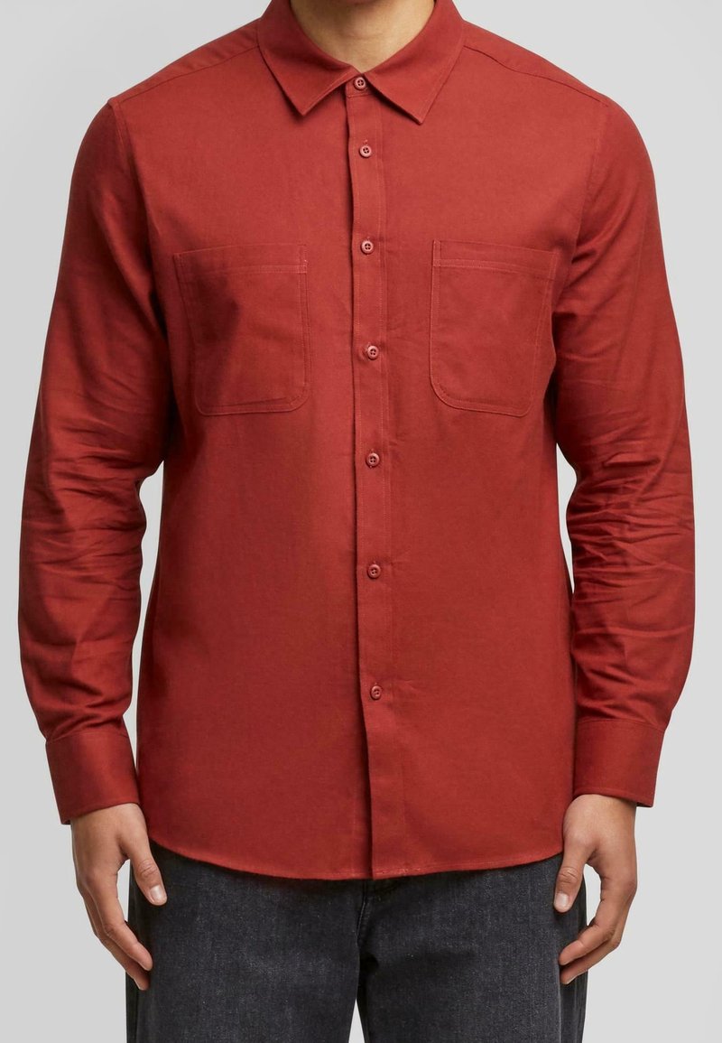 Homme portant une chemise rouge à manches longues avec deux poches poitrine, debout devant un fond gris clair uni.